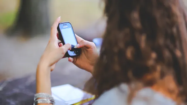 A picture of a woman sending a text. The picture is taken from the back of her head, so you cna see her long curly brown hair and her hands holding a cellphone.